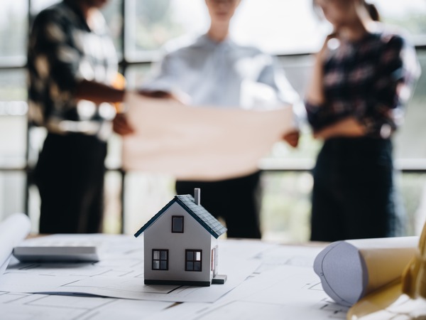 People look at a plan, in the foreground is a desk with rolled and unrolled open plans, with a model of house
